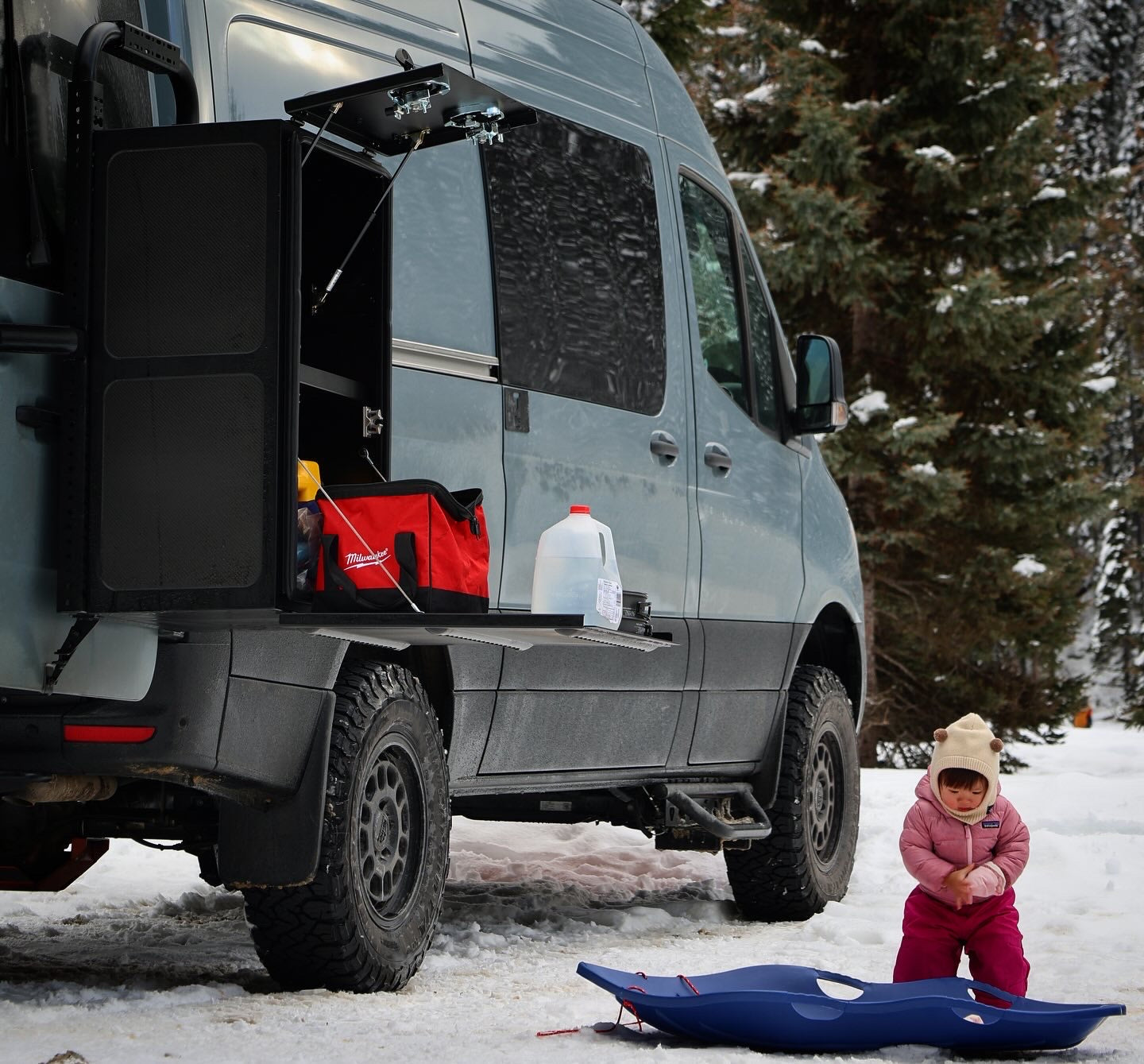 Van with open door in snowy landscape, child with sled nearby